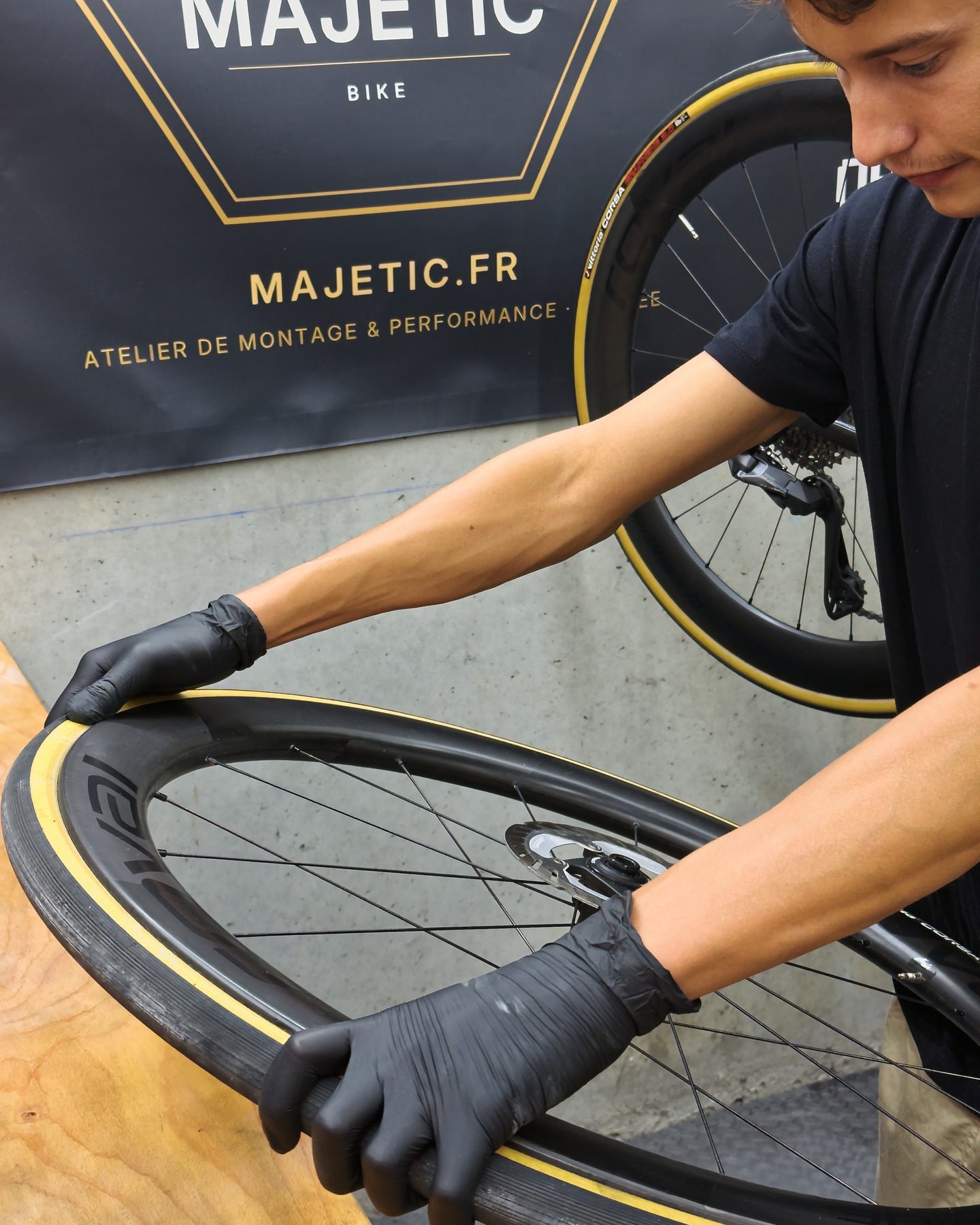 Elian Gaborit, gants d'atelier aux mains, monte un pneu sur une roue à jante carbone posée sur l'établi, devant une bâche Majetic.