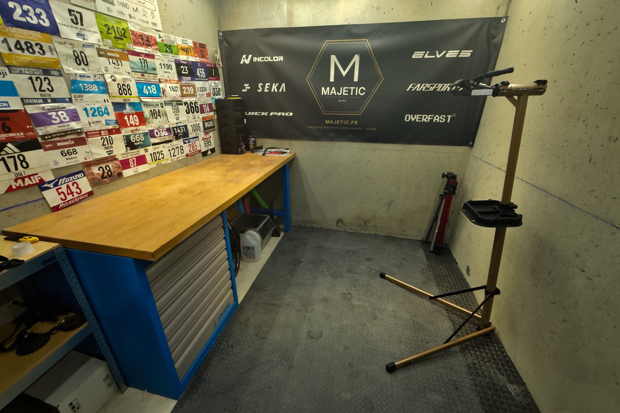 L'établi de l'atelier Majetic, plan de travail en bois sur piètement bleu, mur de dossards de course, bâche des marques partenaires et pied de montage.