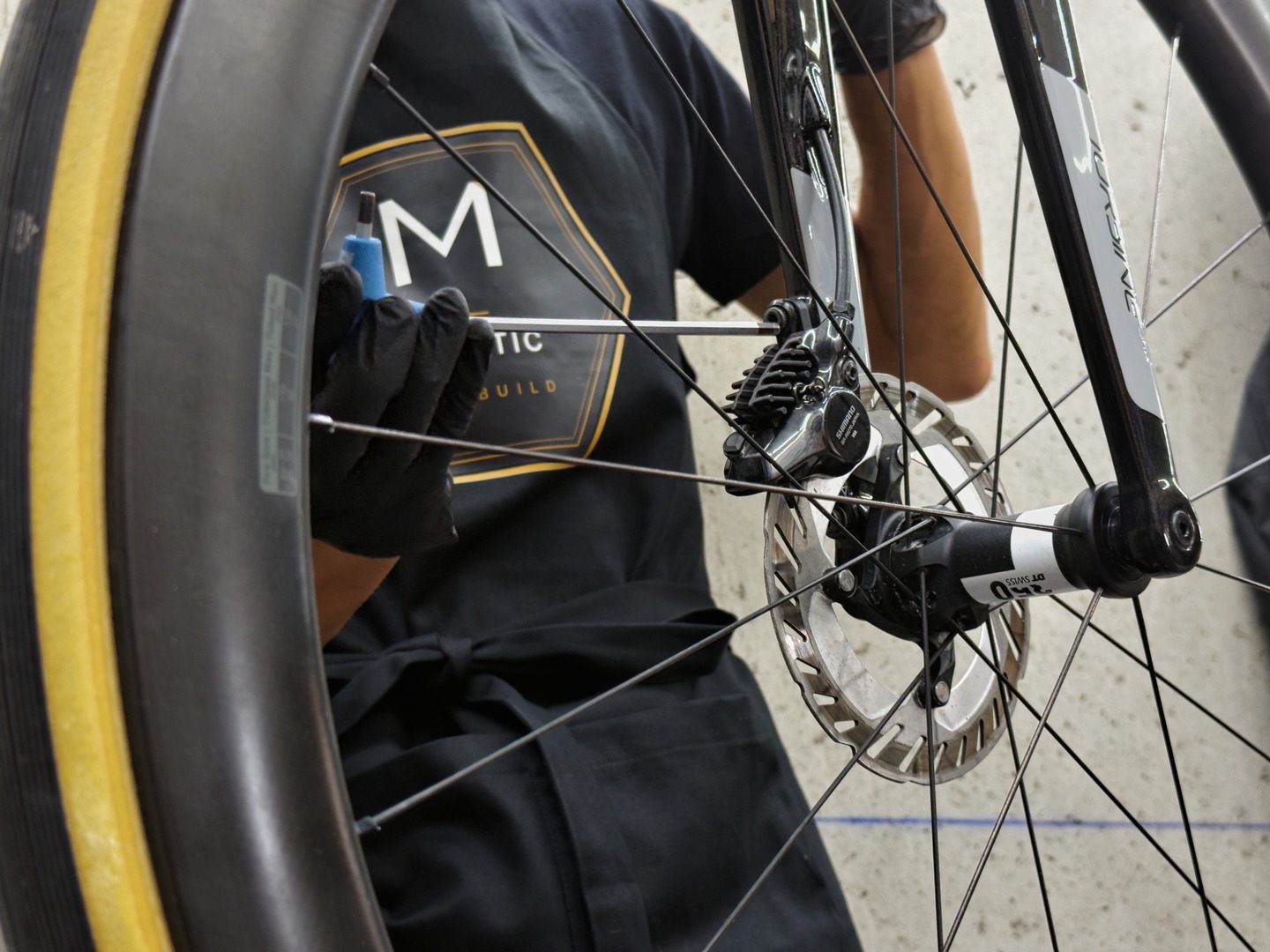 Vue à travers les rayons d'une roue : une main gantée intervient à la clé près du moyeu et du disque de frein d'un vélo de route.