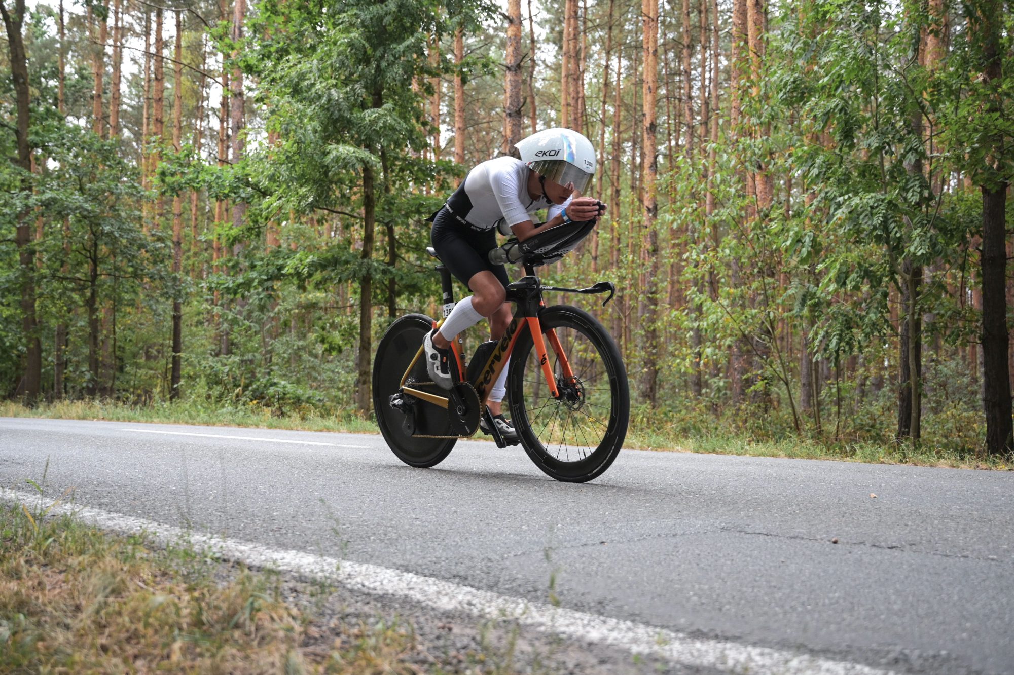 Elian Gaborit de profil, en position aérodynamique sur un vélo de contre-la-montre, casque profilé à visière, roue arrière pleine, sur une route bordée de pins.